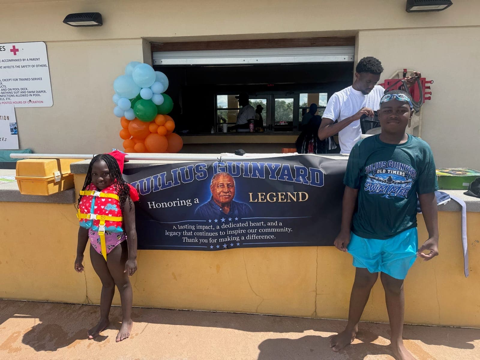 Kids at the pool entrance beside a banner reading “Honoring a Legend” for Julius Guinyard
